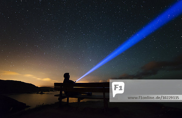 Spanien,  Ortigueira,  Loiba,  Silhouette eines Mannes auf der Bank unter dem Sternenhimmel mit blauem Strahl