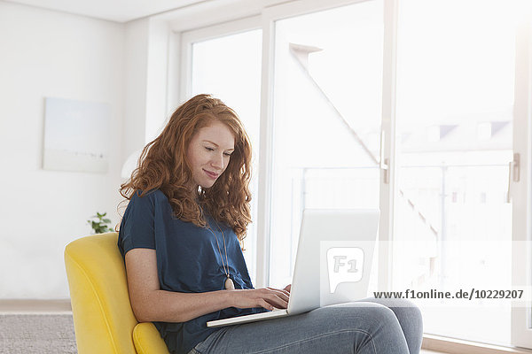 Junge Frau sitzt auf gelbem Sessel in ihrem Wohnzimmer mit Laptop