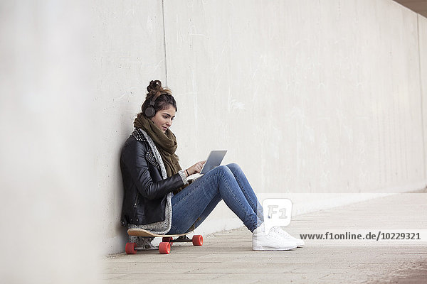 Junge Frau sitzt auf ihrem Longboard mit digitalem Tablett und hört Musik über Kopfhörer.