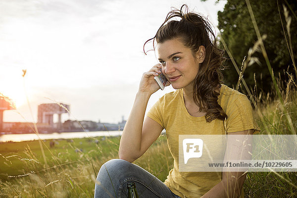 Germany  Cologne  young woman sitting on a meadow near Rhine River telephoning