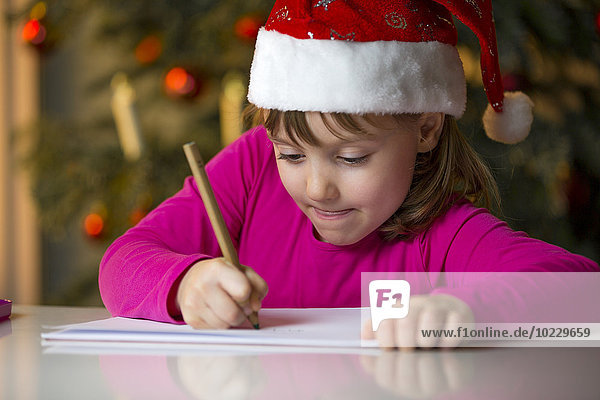 Kleines Mädchen mit Weihnachtsmütze beim Schreiben einer Weihnachtsliste