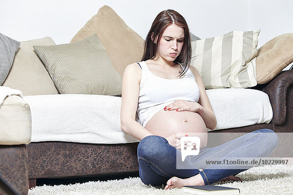 Schwangere Frau sitzt auf dem Boden ihres Wohnzimmers und hält ihren Bauch.