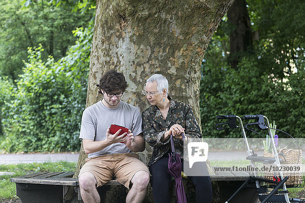Seniorin sieht jungen Mann mit digitalem Tablett auf Parkbank an