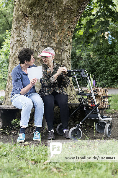 Reife Frau im Gespräch mit älterer Frau mit Rollator auf Parkbank