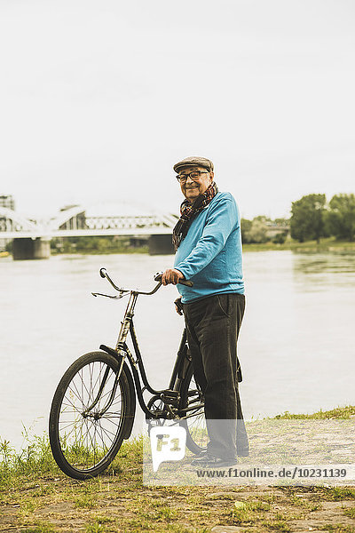 Porträt eines lächelnden älteren Mannes  der mit dem Fahrrad am Wasser steht.