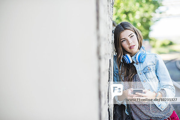 Brunette young woman with headphones and cell phone leaning against wall