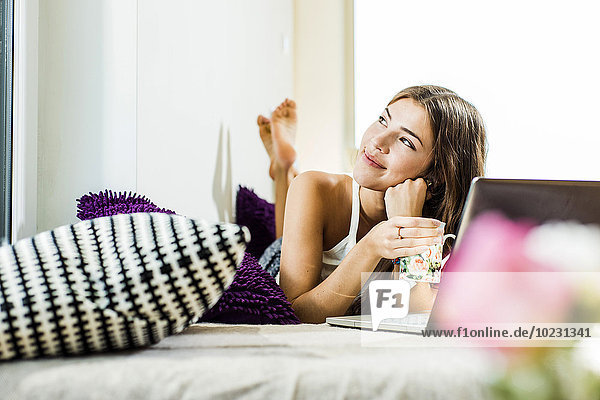 Relaxed young woman lying in bed with laptop looking up