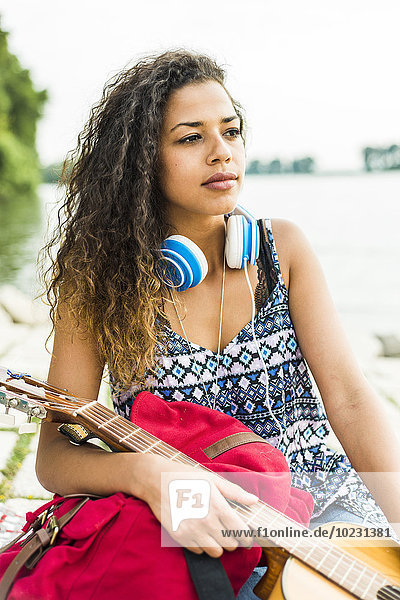 Young woman with headphones  guitar and backpack by the riverside