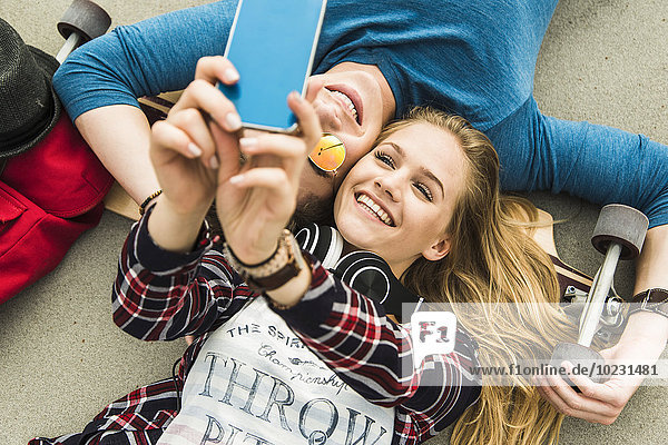 Glückliches junges Paar auf dem Boden liegend mit Skateboard und Blick auf Handy