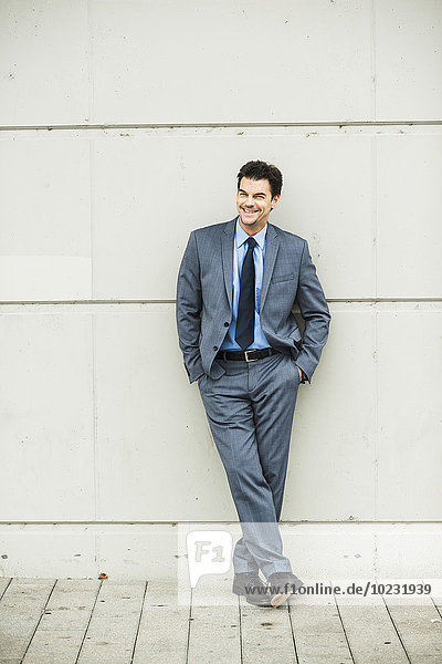 Porträt eines lächelnden Geschäftsmannes im grauen Anzug  der sich an die Betonwand lehnt.