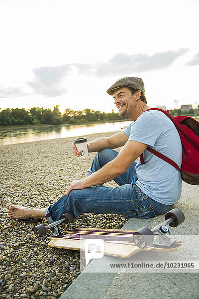 Lächelnder Mann sitzt am steinigen Strand mit Skateboard und Kaffee zum Mitnehmen.