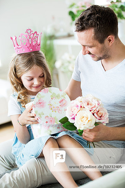 Girl wearing pink crown sitting on father's lap with bunch of flowers