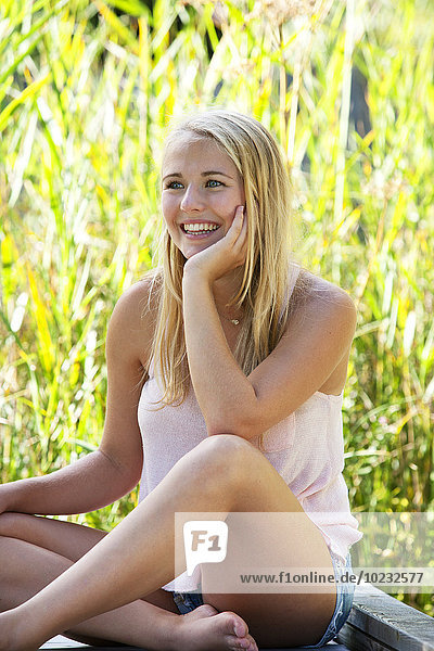 Porträt einer lächelnden jungen Frau  die auf einem Holzsteg sitzt.