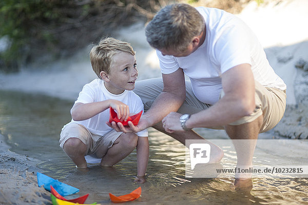 Father and son playing with paper boats at a water pool on a sandy beach
