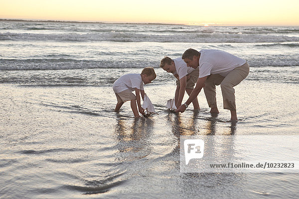 Vater und Söhne beim Spielen mit Spielzeugbooten im Wasser am Strand
