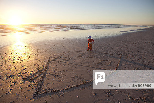 Junge verkleidet als Bauarbeiter mit Bauplänen am Strand mit gezeichnetem Haus im Sand