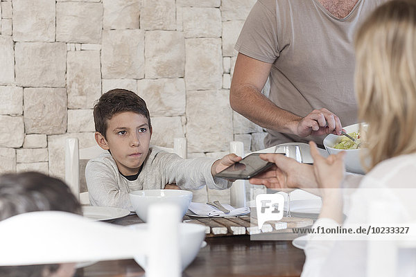 Familie beim Mittagessen am Esstisch mit dem Jungen  der die digitale Tafel der Mutter übergibt.