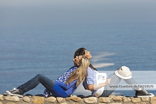Südafrika  zwei glückliche Frauen  die Rücken an Rücken auf einer Wand an der Küste sitzen.