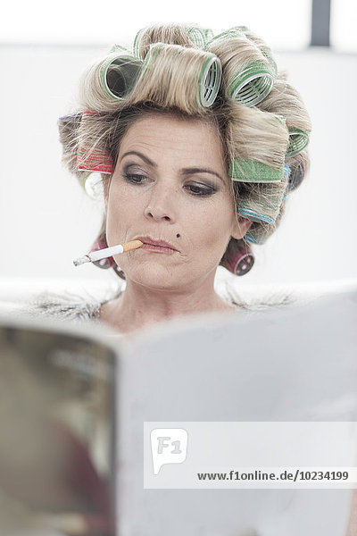 Porträt einer Frau mit Lockenwickler und Zeitschrift  die eine Zigarette raucht.