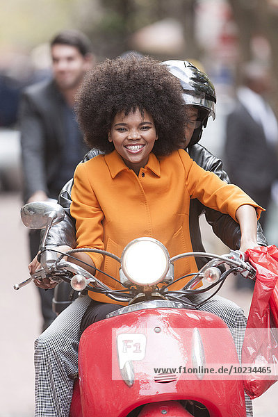 Porträt einer lächelnden Frau  die auf einem Motorroller mit Beifahrer sitzt.