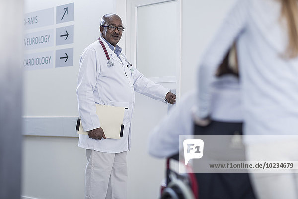 Arzt im Krankenhausflur öffnet Tür und Patient im Rollstuhl