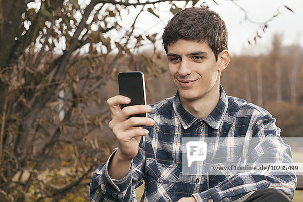 Porträt eines lächelnden jungen Mannes mit seinem Smartphone im Park