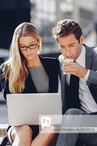 Geschäftsmann und Geschäftsfrau sitzen draußen und arbeiten an einem Laptop.