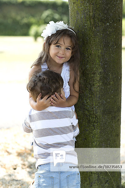 Porträt eines lächelnden Mädchens  das sich an einen Baum lehnt und seinen kleinen Bruder umarmt.