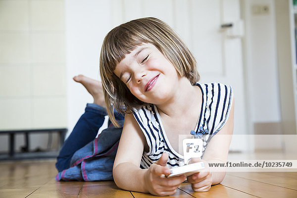Smiling little girl lying on wooden floor with smartphone