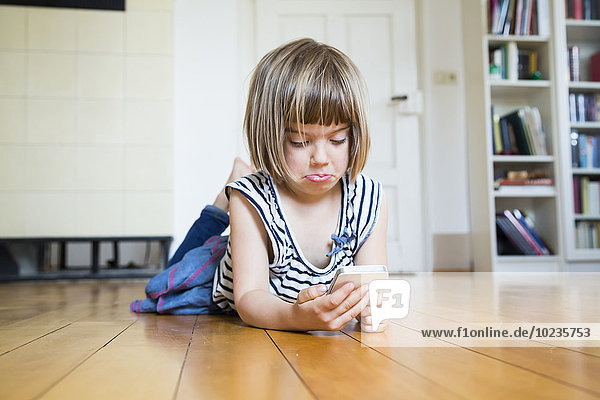 Kleines Mädchen liegt auf Holzboden und sieht skeptisch auf das Smartphone.