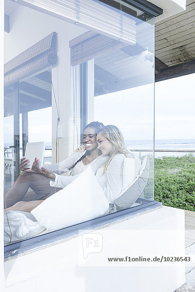 Zwei junge Frauen mit digitalem Tablett entspannen im Strandhaus