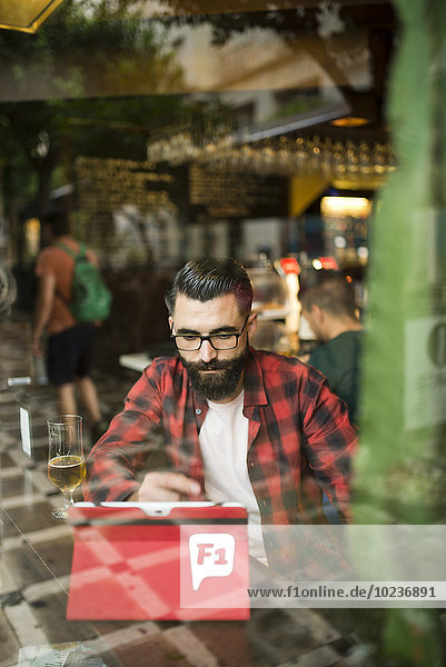 Adult bearded hipster style man touching tablet in bar and drinking alchohol near to window