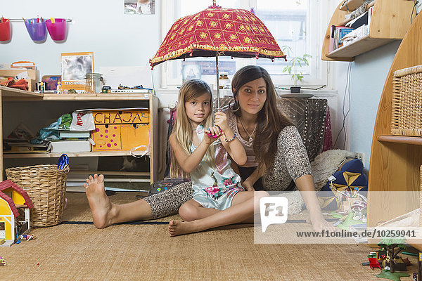 Porträt einer glücklichen Mutter mit einer Tochter  die einen Regenschirm hält  während sie auf einem Hartholzboden im Haushalt sitzt.