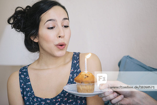 Young woman blowing out candle on cupcake held by man at home