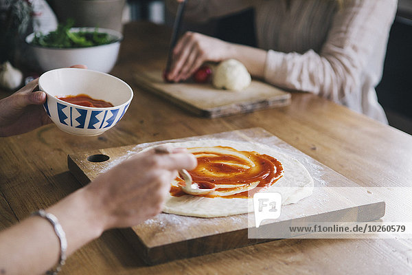 Abgeschnittenes Bild einer Frau  die Tomatensauce über Pizzateig streicht.