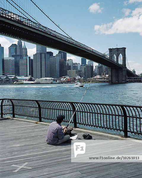 Rückansicht des sitzenden Mannes mit Brooklyn Bridge und Wolkenkratzern im Hintergrund