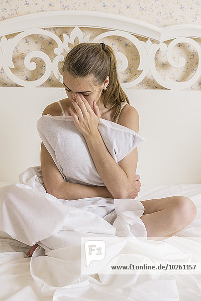 Traurige junge Frau umarmt Kissen während sie auf dem Bett sitzt