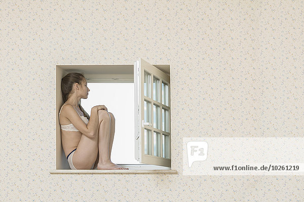 Nachdenkliche junge Frau sitzt auf dem Fensterbrett und schaut weg.