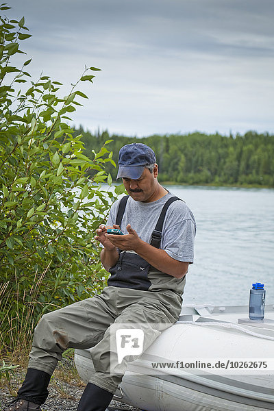 A Alaskan Native man using a smart phone while sitting on a rubber boat along the Kenai River shore  Kenai Wildlife Refuge  Southcentral Alaska  Summer