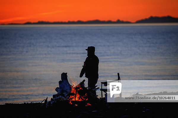 Man enjoying a campfire on the beach during deep red sunset  Homer Spit  Kenai Peninsula  Southcentral Alaska  Spring