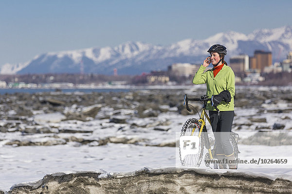 Young woman with her bicycle talks on a cell phone while standing on frozen ice chunks along the Tony Knowles Coastal Trail  Anchorage  Southcentral Alaska