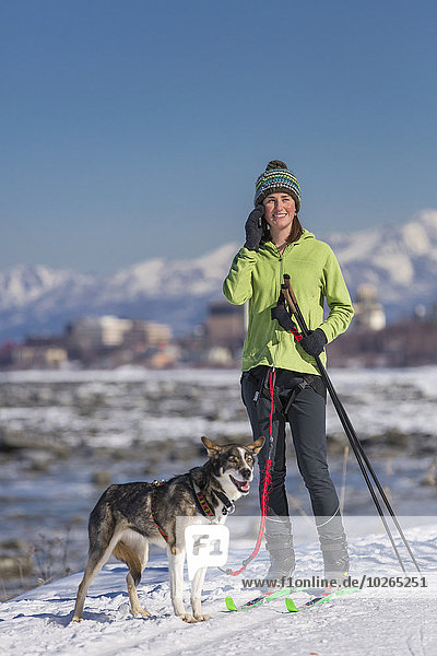 A young woman takes a break from skijoring with her Alaska Husky sled dog to talk on her phone  Tony Knowles Coastal Trail  Anchorage  Southcentral Alaska  USA.