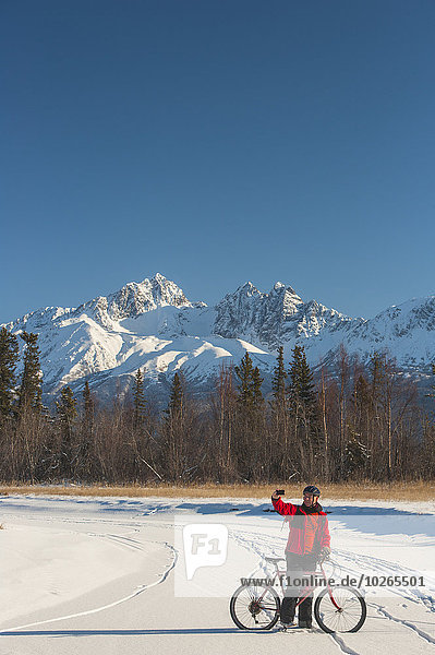 Man bicycling on frozen Rabbit Creek in the Palmer Haystack Game Refuge takes a selfie with Twin Peaks in the background  Southcentral Alaska  winter