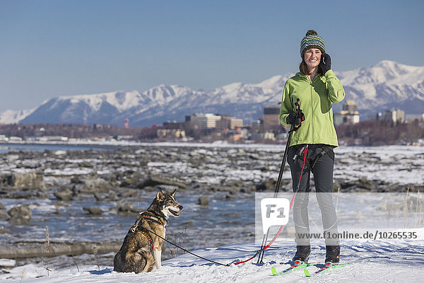 A young woman stops and talks on a cell phone while she skijors with her Alaska Husky sled dog down the Tony Knowles Coastal Trail  Anchorage  Southcentral Alaska  USA.