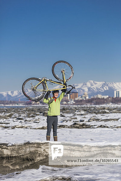 A young woman holds her bicycle over her head in triumph while standing on ice next to the Tony Knowles Coastal Trail  Anchorage  Southcentral Alaska  USA.