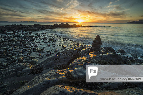 Woman sits on the shore of the Pacific Ocean watching the sunset near Jordan River; Vancouver Island  British Columbia  Canada