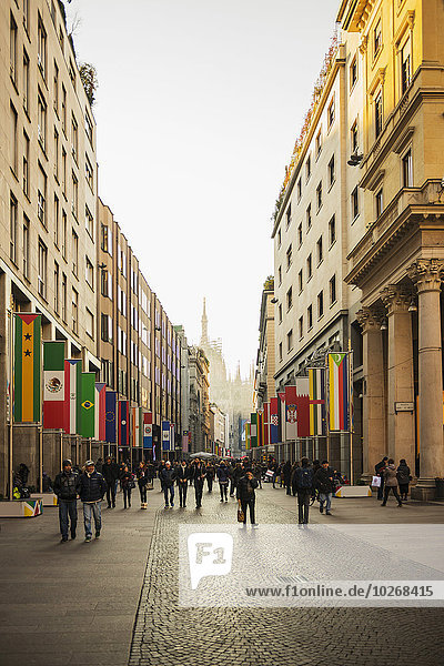 Fußgänger gehen auf einer Straße  die mit internationalen Flaggen auf den Gebäuden und der Mailänder Kathedrale in der Ferne gesäumt ist. Mailand  Lombardei  Italien