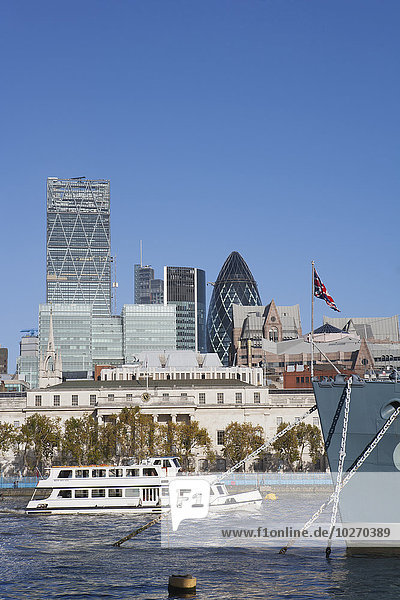 A tourist boat passes the prow of the warship HMS Belfast  docked on the River Thames and part of the Imperial War Museum  with skyscrapers in the City of London  including The Gherkin by Norman Foster; London  England