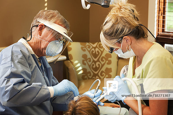 A dentist and his assistant wearing protective gear and doing a dental proceedure on a patient; United States of America