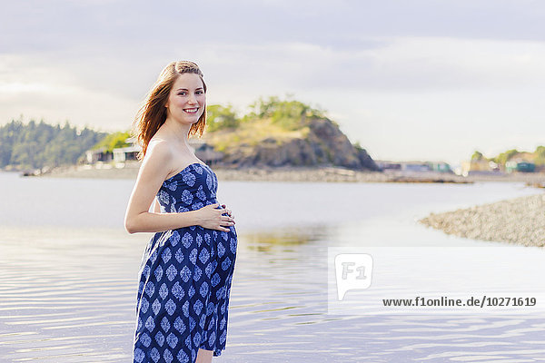 'A young woman poses with a smile for a pregnancy maternity portrait on a beach at the ocean; Nanaimo  British Columbia  Canada'